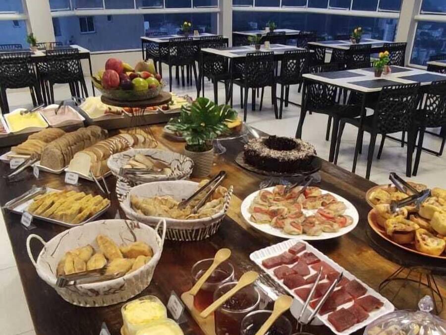 Un montaje tipo bufé con diversos alimentos y utensilios, dispuesto sobre una larga mesa en una sala con ventanas amplias y sillas negras.