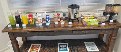 A wooden table with various food and drink items, including a coffee maker, jars of nuts, and boxes of granola.