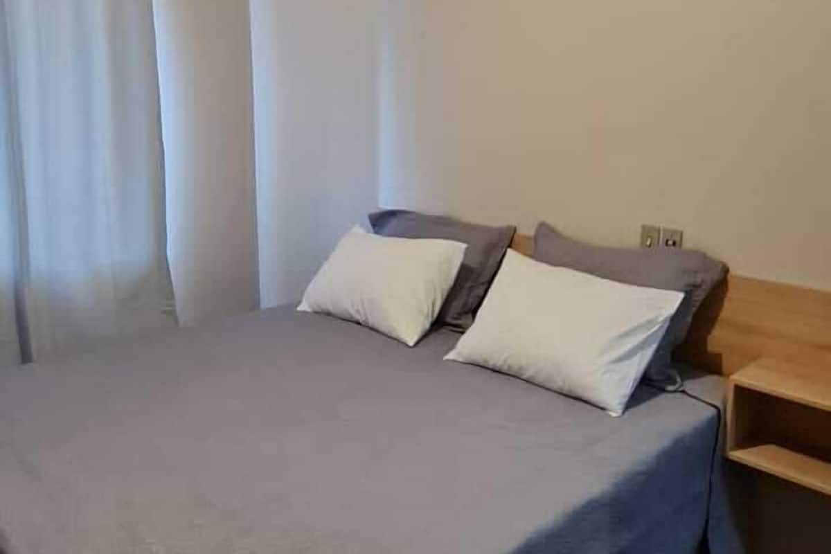A neatly made bed with a grey bedspread and two pillows, a wooden floor, and a ceiling light fixture.