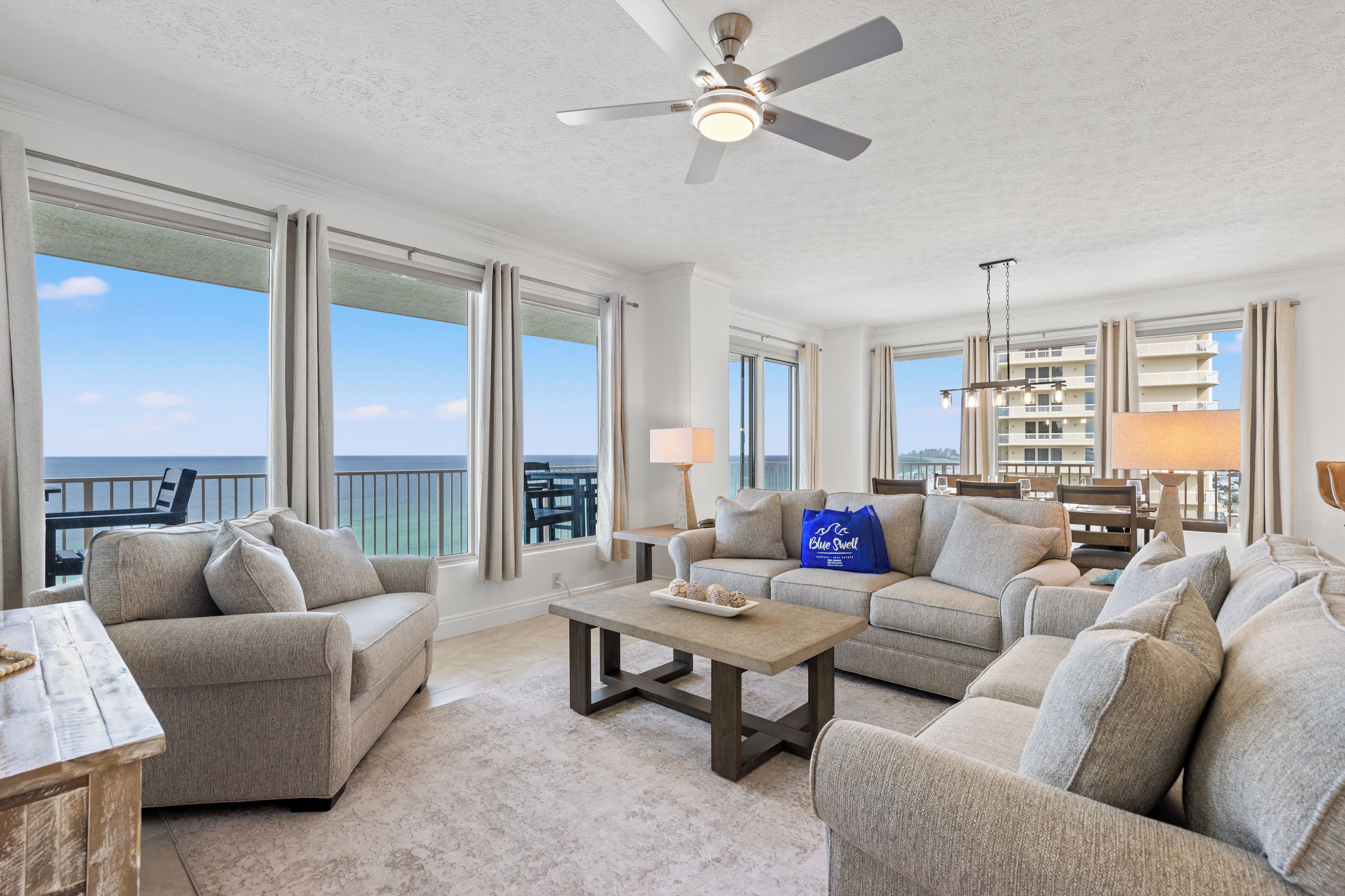A spacious living room with a large window overlooking the ocean, a ceiling fan, and a seating area with a coffee table.