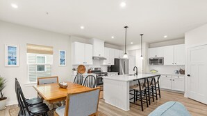 A modern kitchen with white cabinetry, a central island with bar stools, a dining area with a wooden table and chairs, and pendant lighting.