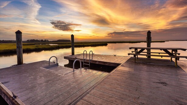A wooden dock with a metal ladder and a picnic table, overlooking a calm body of water during sunset.