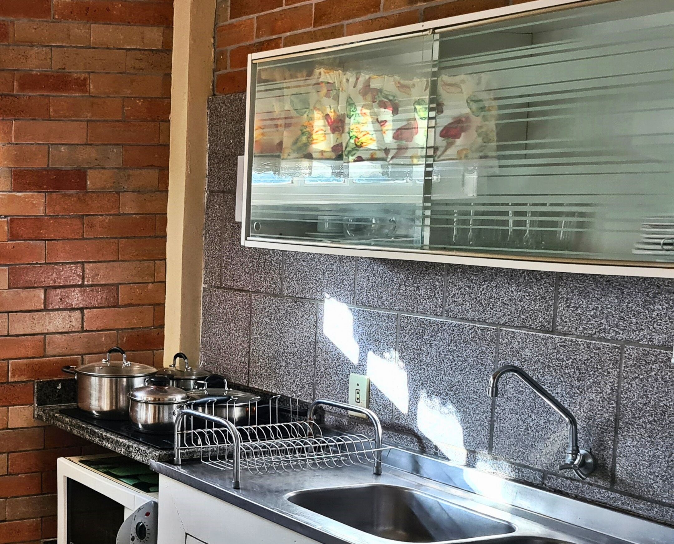 A kitchen with a brick wall, granite backsplash, stainless steel sink, and a dish rack.