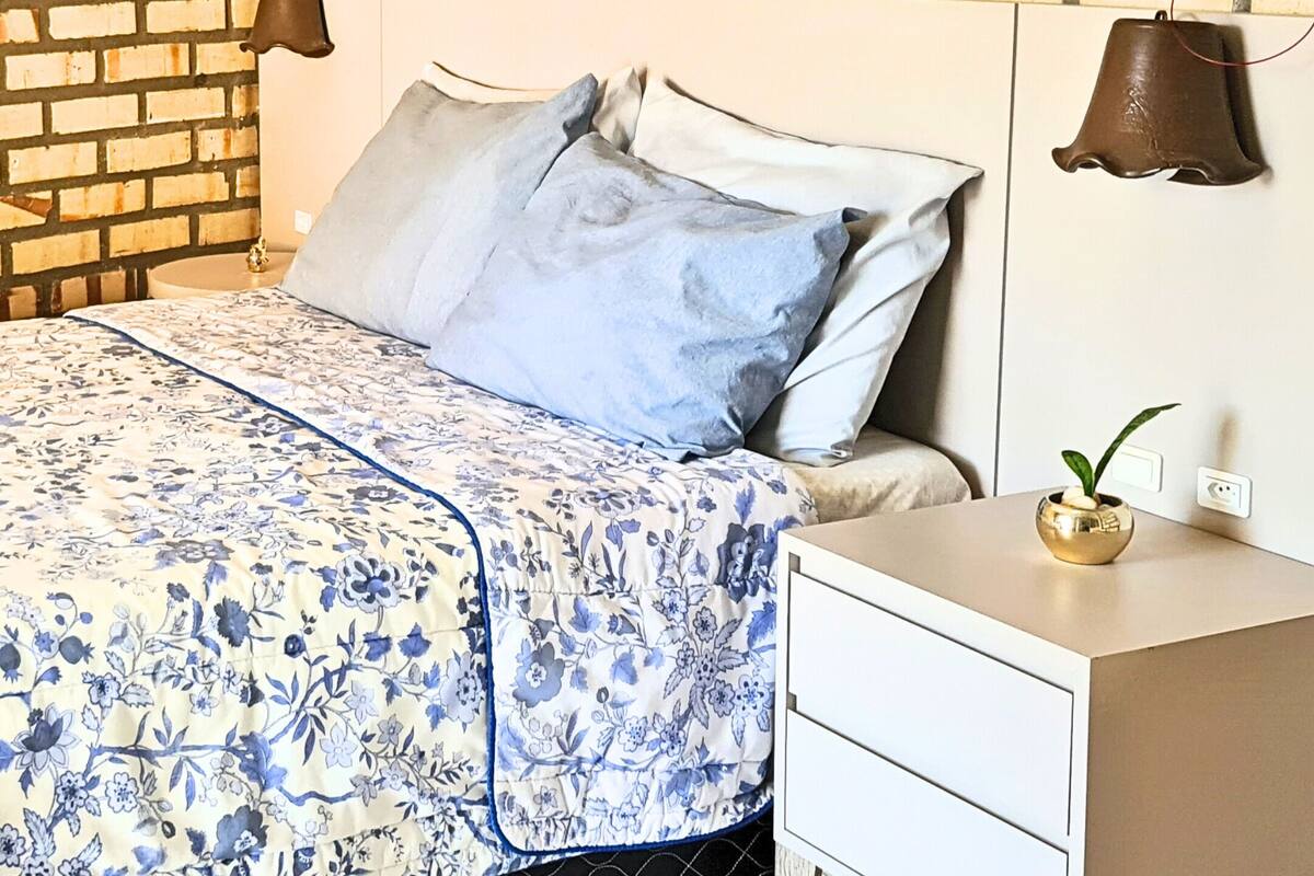 A bedroom with a brick wall, a bed with floral bedding, two bedside lamps, and a wall-mounted painting.
