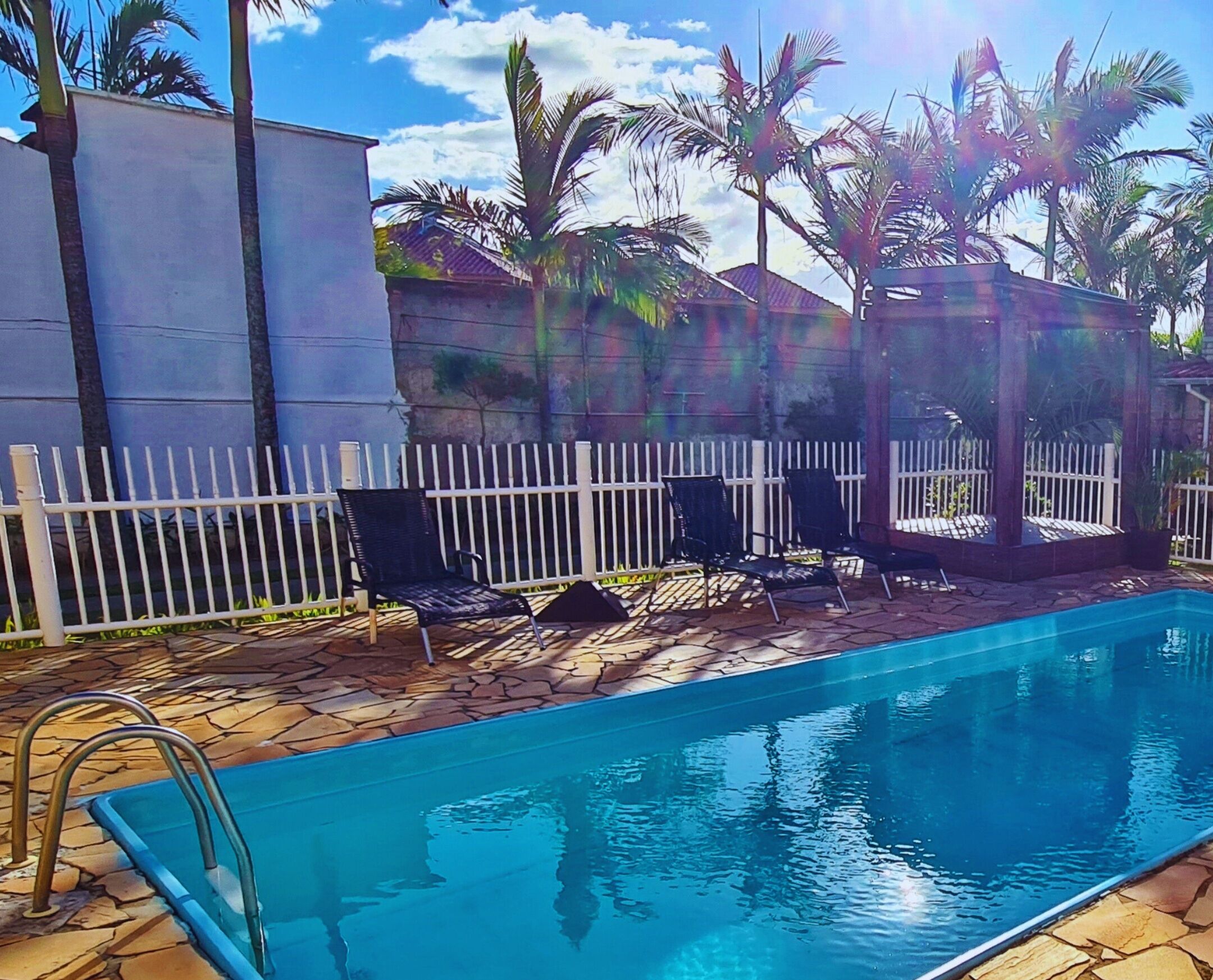 A swimming pool with a white fence, lounge chairs, and palm trees in the background.