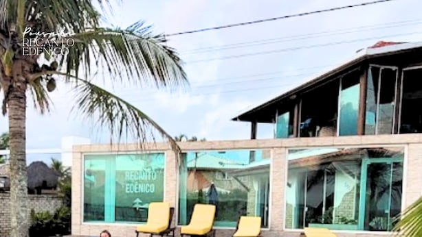 Een gebouw aan het strand met een bord waarop 'RECANTO EDENICO' staat. Er staan gele ligstoelen op een houten terras.