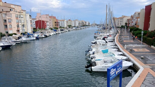 Un port de plaisance où des bateaux sont amarrés le long d’un canal, entouré d’immeubles à plusieurs étages.