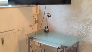 Une pièce équipée d’un téléviseur à écran plat fixé au mur, d’un bureau avec un plateau en verre et une lampe, et d’un radiateur posé sur le sol.