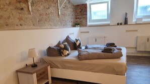 Une chambre avec un plancher en bois, un lit blanc agrémenté de plusieurs oreillers, une table de chevet avec une lampe, un mur en briques et une fenêtre donnant sur un paysage agréable.