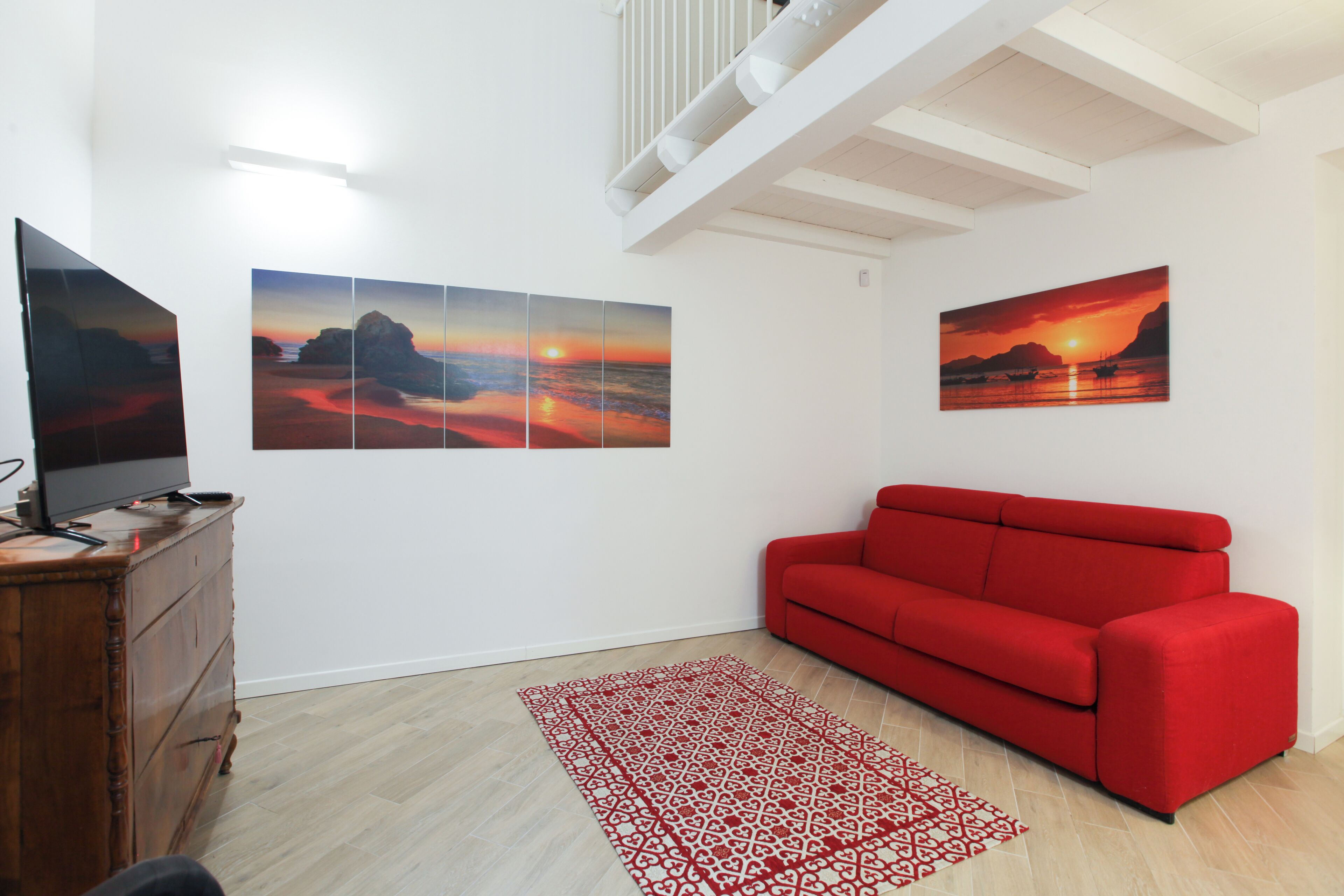 Ein Wohnzimmer mit einem roten Sofa, einer Holzkommode und drei Sonnenuntergangsbildern an der Wand.