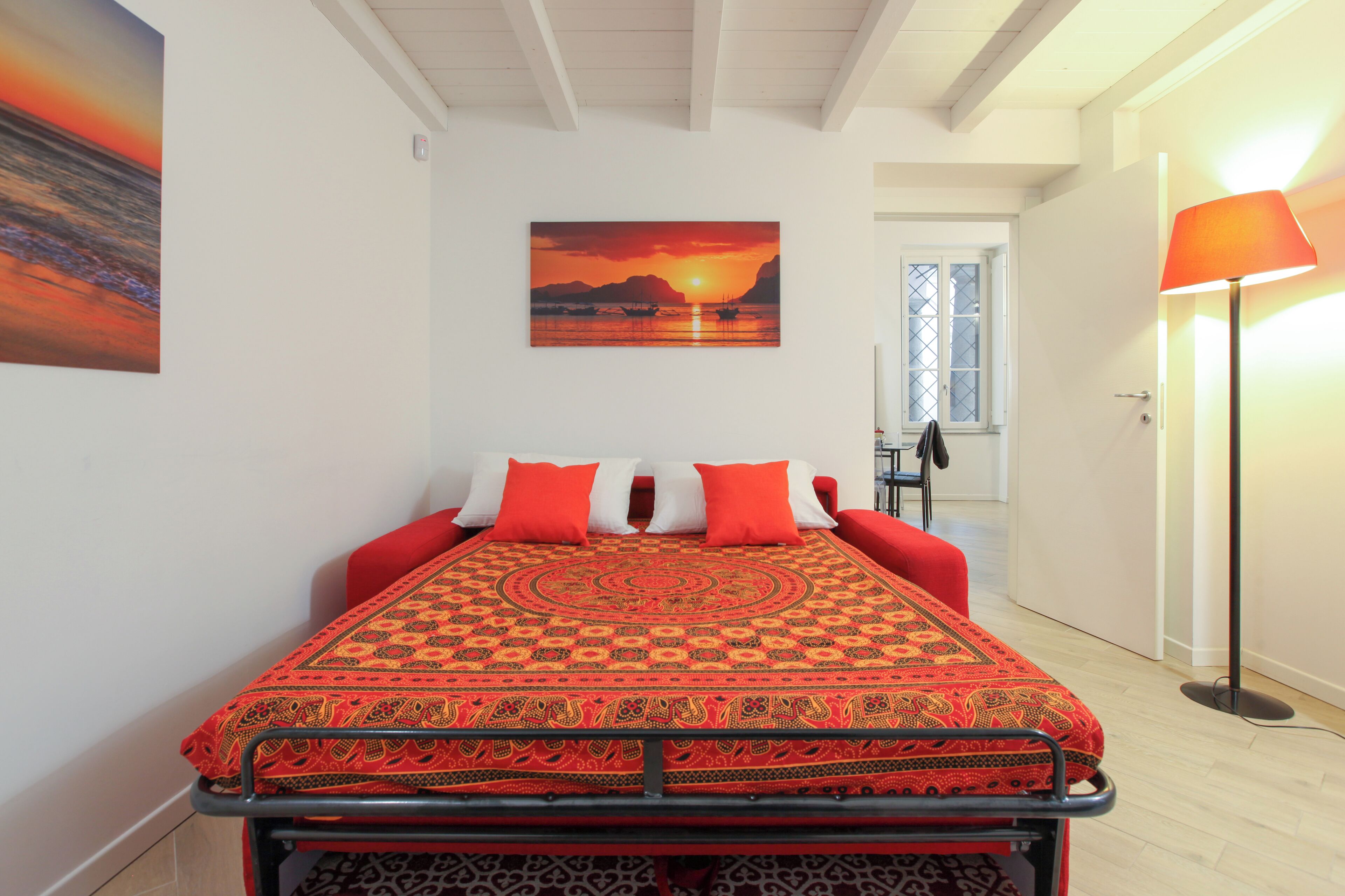 Een slaapkamer met een bed, rode en witte kussens, een beddengoed met een patroon, een schilderij van een zonsondergang, en een vloerlamp.