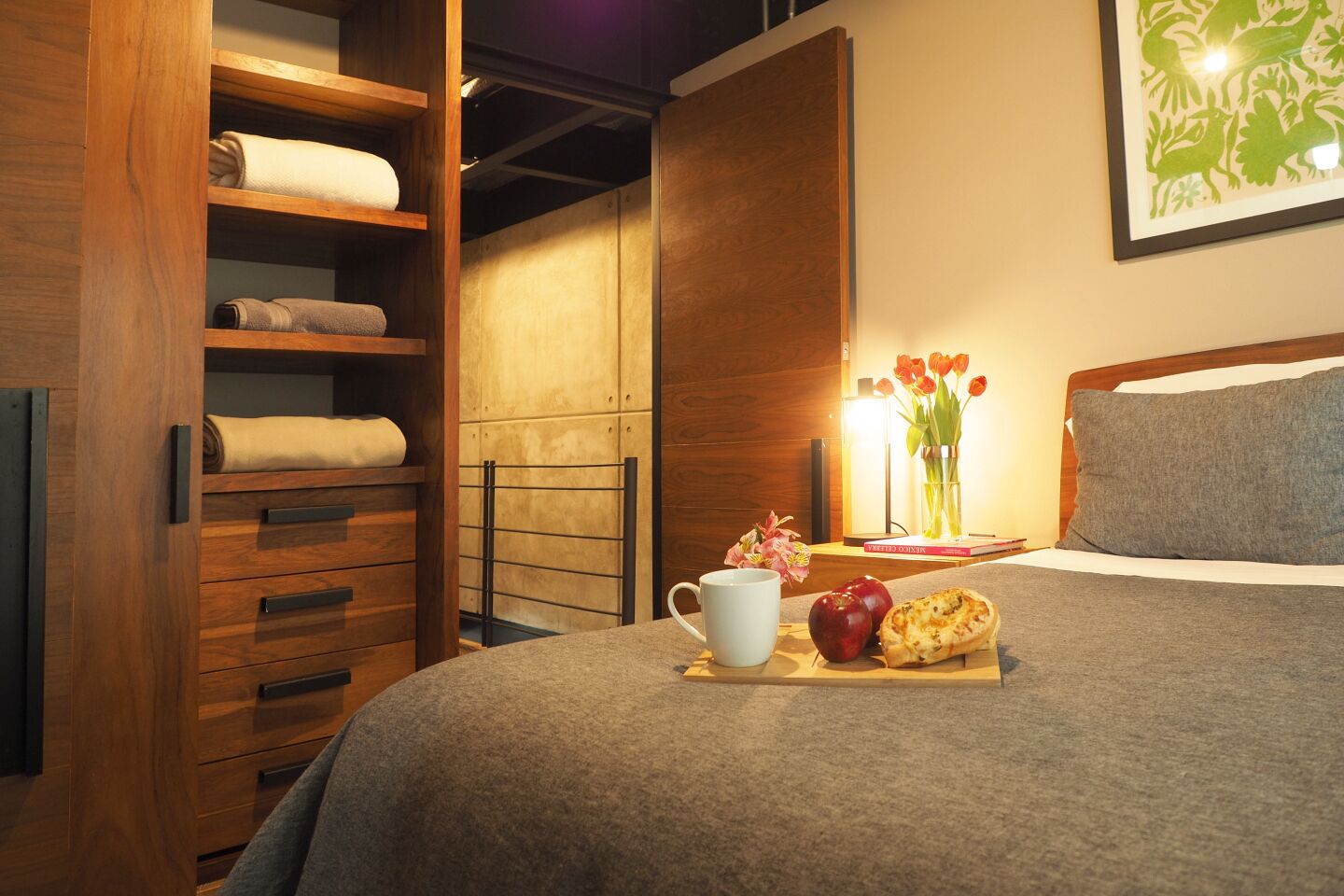 A neatly made bed with a grey blanket, a bedside table with a book, a vase of tulips, and a tray with an apple and a pastry.
