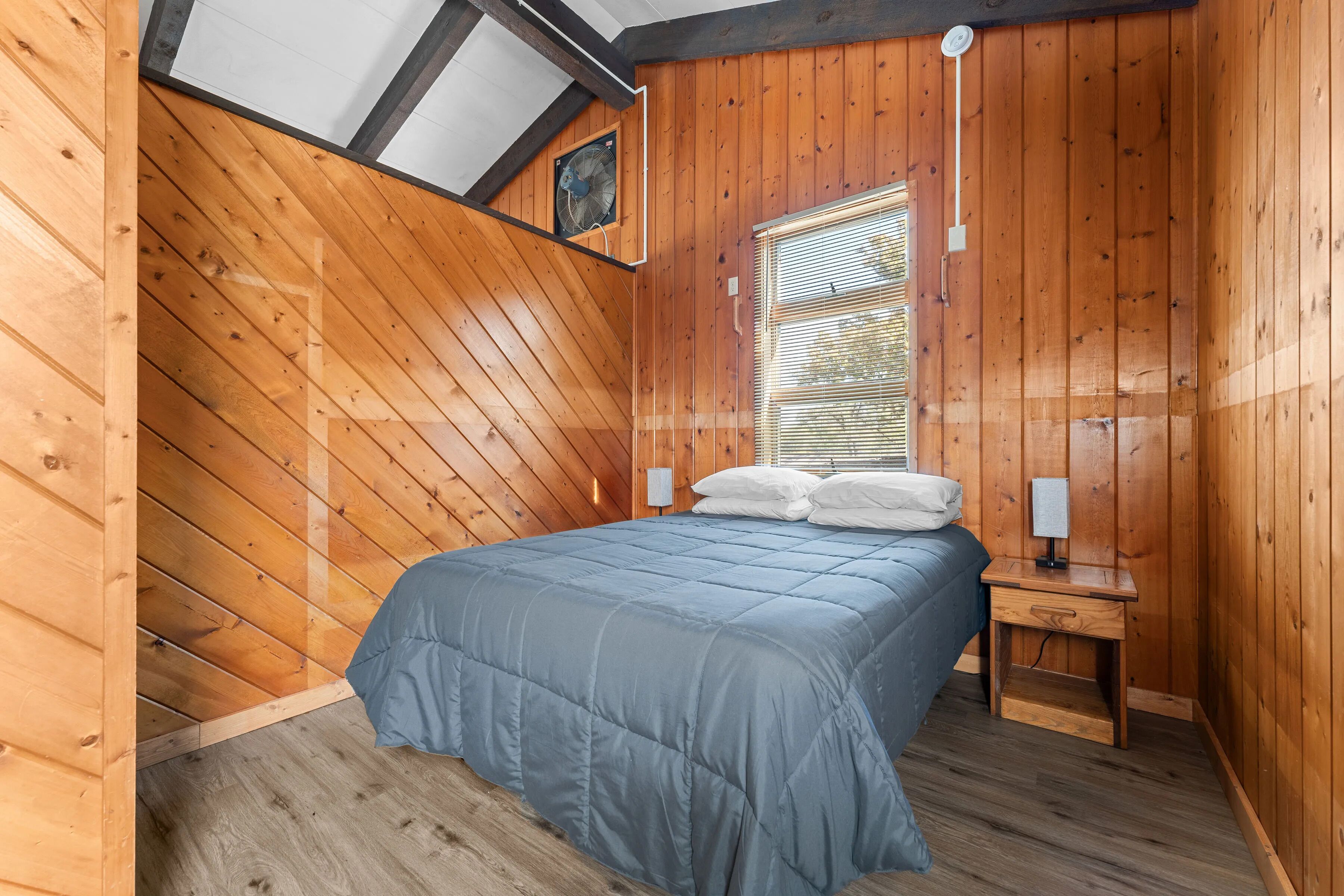A bedroom with a sloped ceiling, wooden walls, a bed with a blue bedspread, a bedside table with a lamp, and a window with blinds.