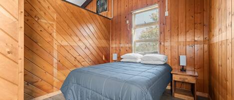 A bedroom with a sloped ceiling, wooden walls, a bed with a blue bedspread, a bedside table with a lamp, and a window with blinds.