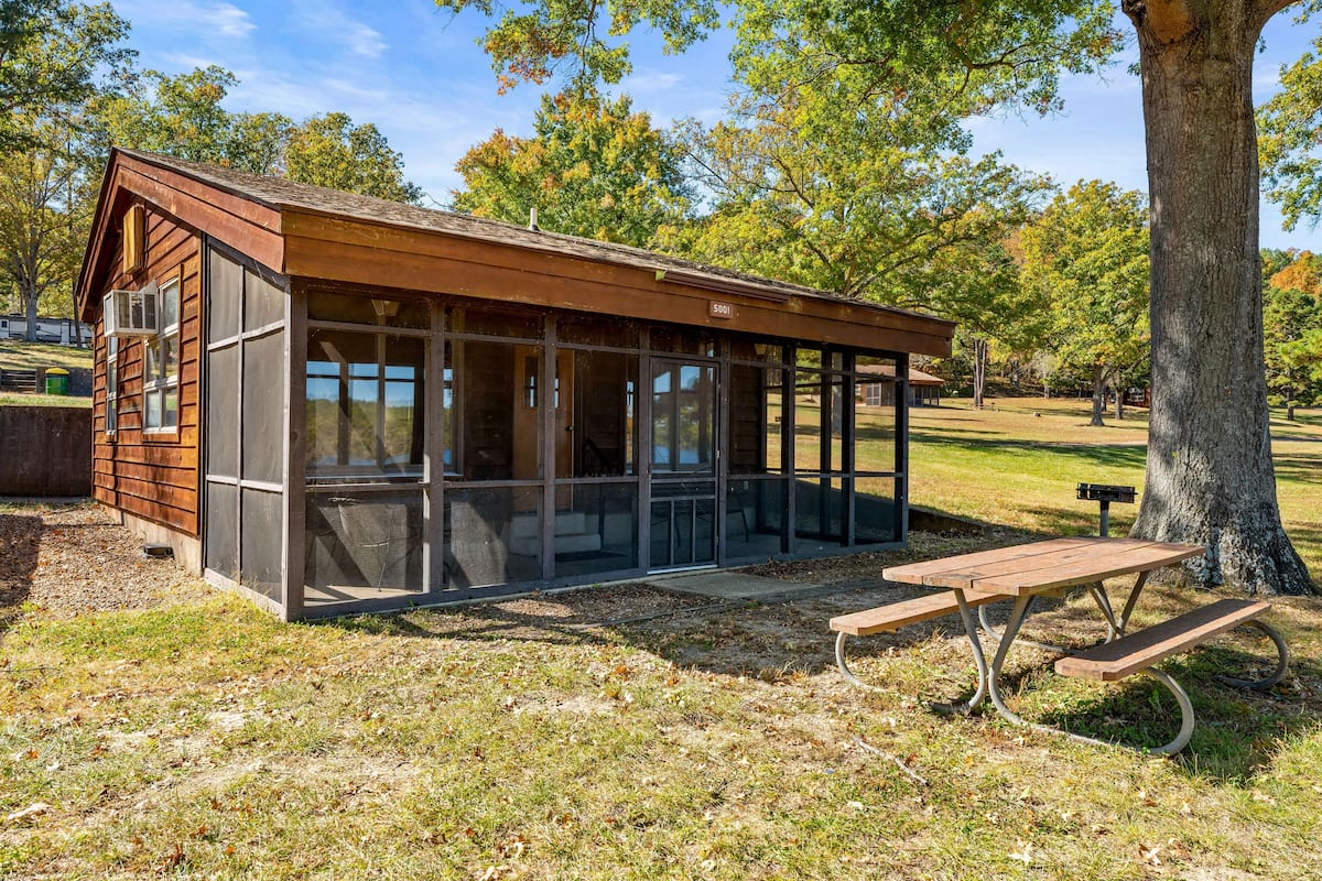 Een klein houten gebouwtje met grote ramen, een picknicktafel en een bank.