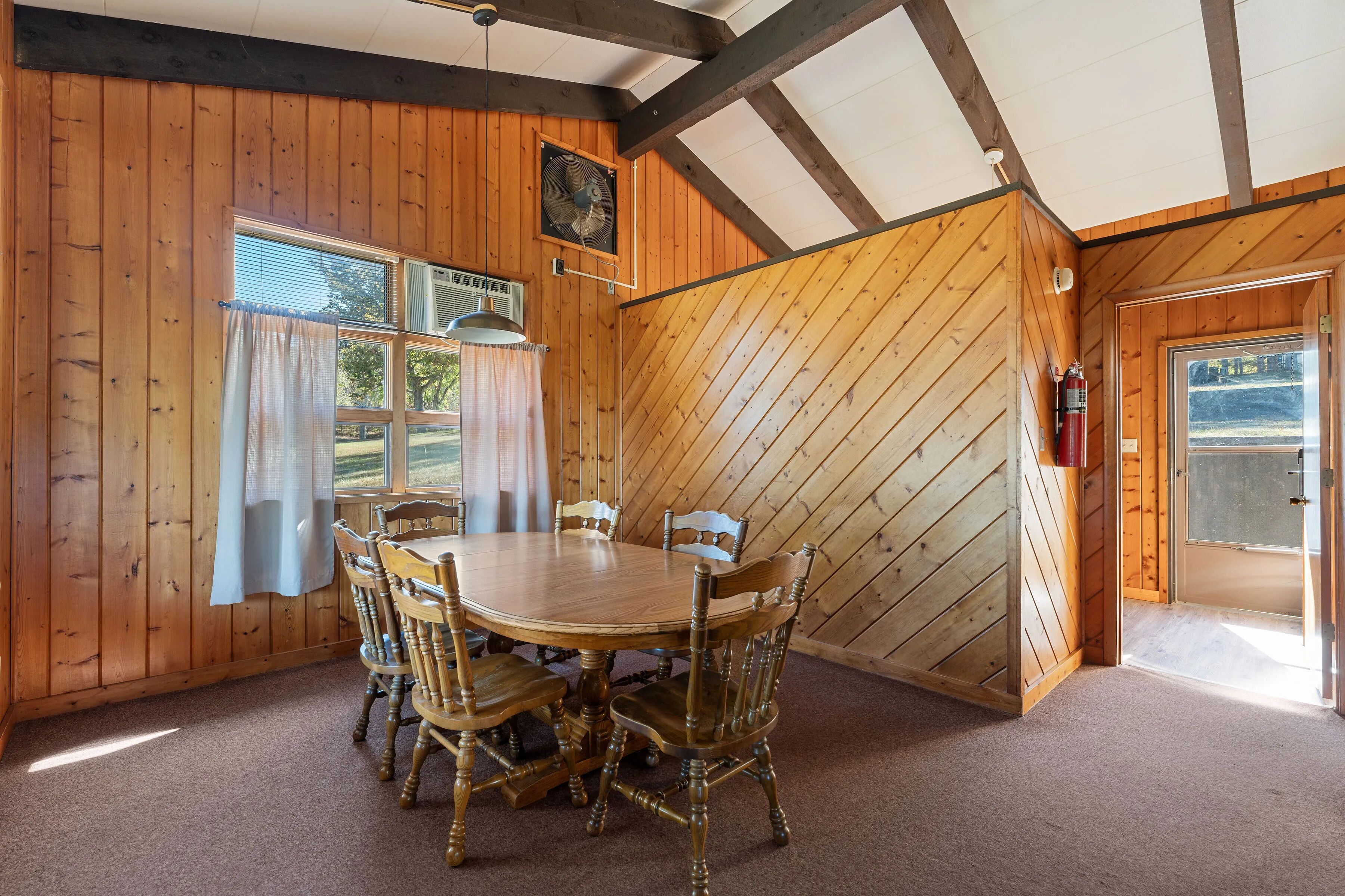 A wooden-paneled room with a round wooden table and six chairs, a ceiling fan, and a fire extinguisher near the door.