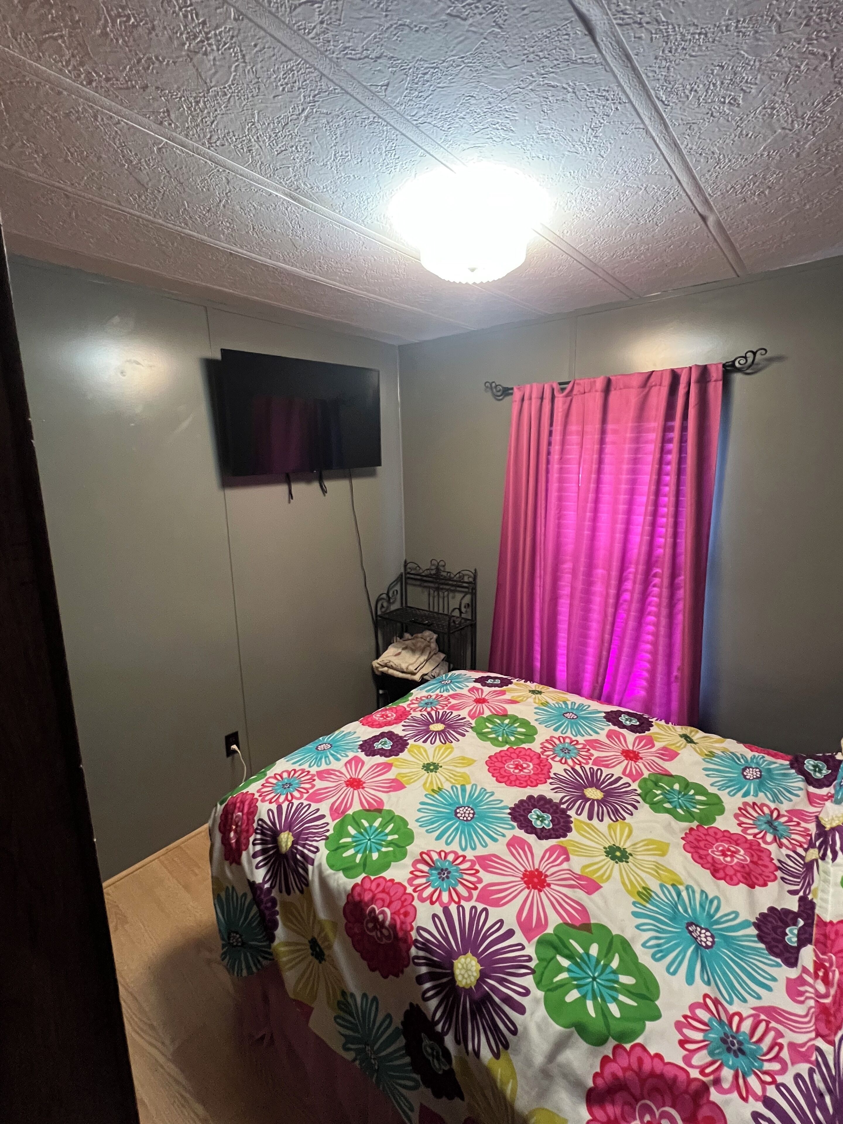 Een slaapkamer met een bloemenbedekker, een televisie aan de muur en een metalen nachtkastje met een opgevouwen deken.