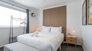 A modern hotel room with a large bed, a bedside table, a nightstand, a window with curtains, and a wall-mounted plant.