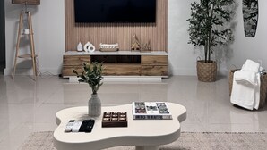 A modern living room with a large flat-screen TV mounted on a wooden panel wall, a white coffee table with a vase and remote controls, a floor lamp, and a potted plant. - Charming 1-bedroom apartment in North Bay Village with WiFi, AC (North Bay Village)