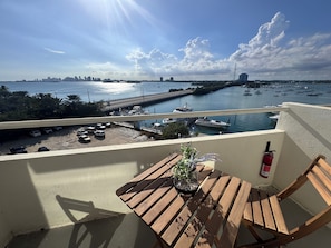 Un balcón con mesa y sillas de madera, con vistas a una marina con barcos y el perfil de la ciudad.