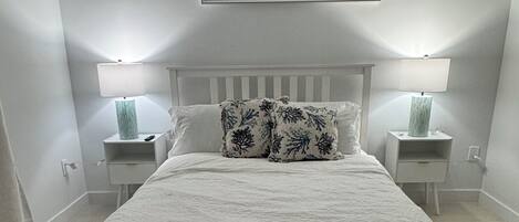 A bedroom with a large bed, two bedside tables with lamps, a framed picture above the bed, and a white headboard.