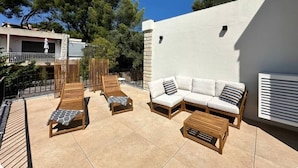 A patio with wooden loungers, a sofa, and a table, surrounded by a white wall and trees.