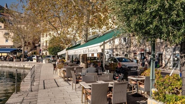 An outdoor café with tables and chairs, a stone pathway, and a body of water. - Antigona (Bakar)