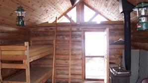 Une cabane en rondins avec un lit en mezzanine, une échelle en bois, un poêle et une grande fenêtre.