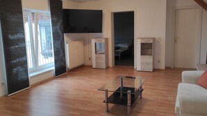 Un salon moderne avec une table basse en verre, un canapé blanc agrémenté de coussins, un téléviseur à écran plat fixé au mur et un plancher en bois.