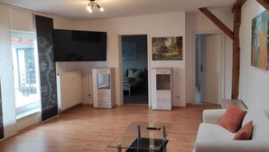 Un vaste espace de vie avec un canapé blanc, une table basse en verre et un téléviseur fixé au mur. Le plafond est orné de poutres en bois et un tableau est accroché au mur.