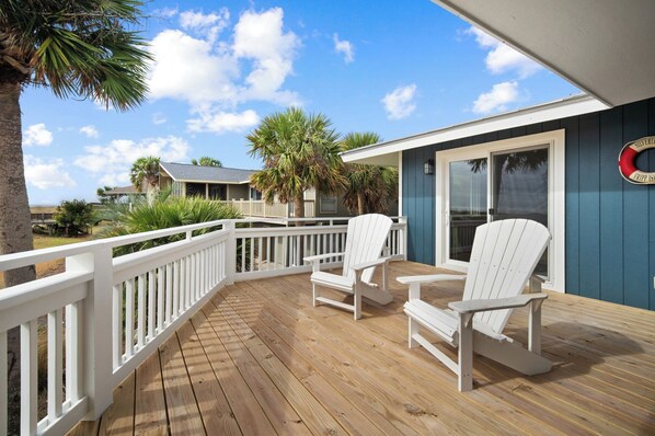 A wooden deck with white Adirondack chairs, a blue building with a lifebuoy, and a palm tree. - Beachfront Home, Oceanfront, Dog Friendly - 710 Silverside (Fripp Island)