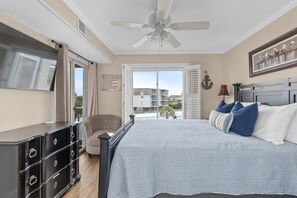 A bedroom with a bed, a dresser, a chair, a ceiling fan, and a view of buildings outside.