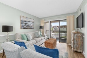 A living room with a sofa, armchairs, a coffee table, a television, and a sliding glass door leading to a balcony.