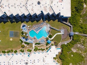 An aerial view of a resort with a large swimming pool, multiple lounge areas, and a bridge connecting different sections.