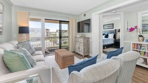 A living room with a sofa, a small table, a bookshelf, and a view of the outside. - A Place at the Beach - 2 bedroom, 2 baths - Pools, Waterslide, Mini-golf & more! (Atlantic Beach)