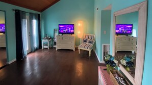 Ein Zimmer mit Türkiswänden, Holzboden, einem Fernseher auf einer Kommode, einem Stuhl, einer Lampe und einem Spiegel.