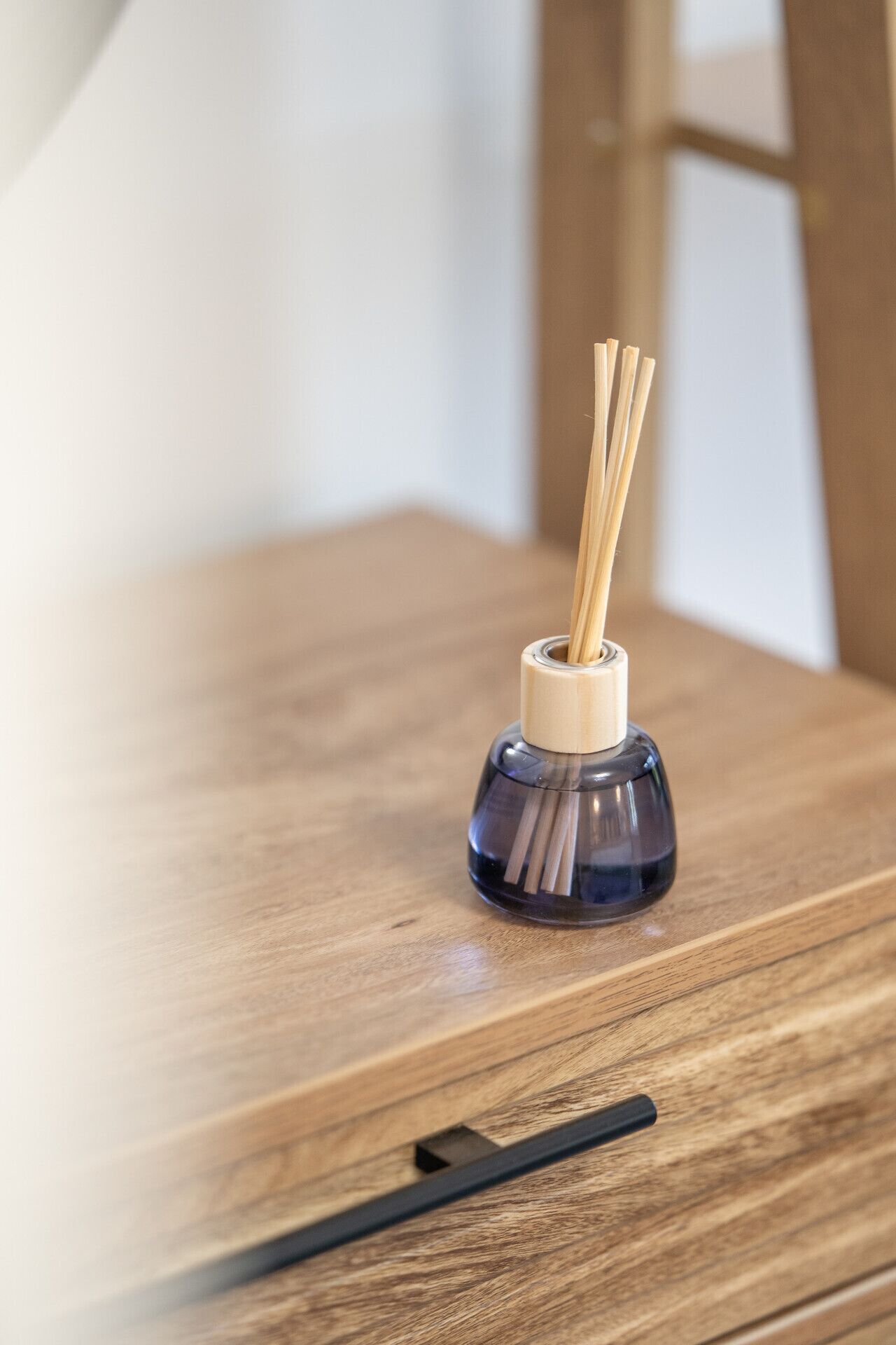 A dark blue glass bottle with a wooden lid and reed diffuser sticks on a wooden table.