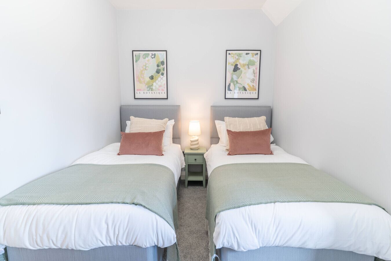 Une chambre double avec deux lits, chacun équipé d’un oreiller et d’une couverture. Une table de chevet avec une lampe se trouve entre les deux lits. Deux reproductions encadrées de motifs botaniques sont accrochées au mur, au-dessus des lits.