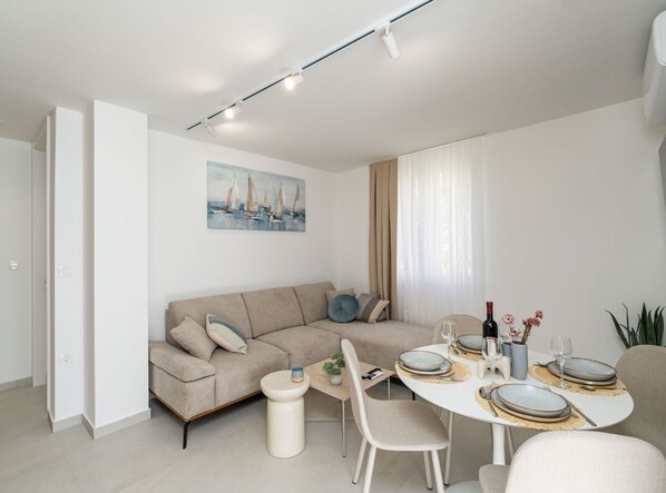 Una sala de estar moderna con un sofá beige, una mesa de comedor preparada para cuatro personas y un cuadro de veleros en la pared.