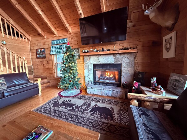Interior acogedor de una cabaña de troncos con chimenea, un árbol de Navidad, una cabeza de venado montada en la pared y un televisor también colgado.