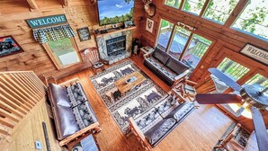 A cozy cabin interior with a fireplace, wooden furniture, and a large window offering a view of the outdoors. - A Bear’s Eye View – Epic 4BR 3BA Cabin wGame Room, Hot Tub & Stunning Vistas!  (Sevierville)