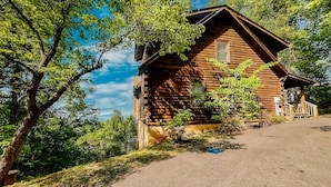 A rustic log cabin with a porch, surrounded by lush greenery and trees. - A Bear’s Eye View – Epic 4BR 3BA Cabin wGame Room, Hot Tub & Stunning Vistas!  (Sevierville)