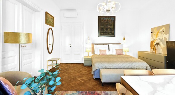 Un dormitorio con cama, mesitas de noche, cómoda, silla, lámpara, espejo y un cuadro en la pared.