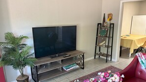 A living room with a flat-screen TV, a wooden entertainment unit, a potted plant, a red sofa, and a dining area with a yellow tablecloth. - Charming 2-bedroom house with WiFi, AC in beautiful Gulf Breeze (Gulf Breeze)