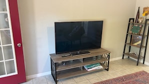 A living room with a TV on a wooden stand, a red door, and a shelf with decorative items. - Charming 2-bedroom house with WiFi, AC in beautiful Gulf Breeze (Gulf Breeze)