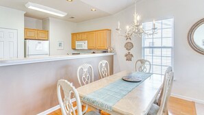 A modern kitchen with wooden cabinets, a white refrigerator, and a dining area with a table and chairs. - 5 Mi to Shore: Condo w/ Patio in Sunset Beach! (Sunset Beach)