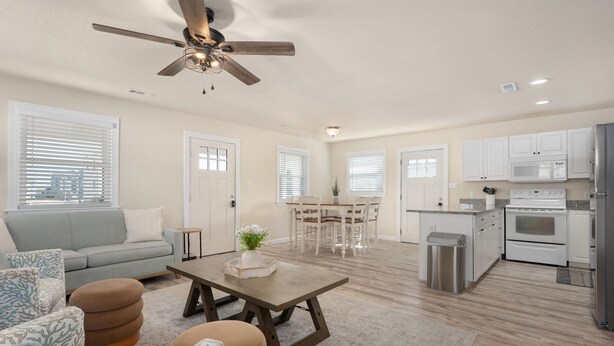 A spacious living room with a gray sofa, a wooden coffee table, and patterned ottomans. A dining area with a white table and chairs is adjacent to the kitchen, which features white cabinets, a microwave, and a stainless steel refrigerator. - Pool/Hot tub -Fenced yard -1 dog friendly (Nags Head)
