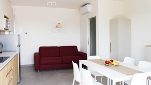 A compact living space with a kitchenette, a red sofa, a dining table with chairs, and a fruit bowl. - Dainese Apartments, Casa Miriam (Jesolo)