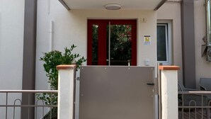Entrada de un edificio con una verja metálica cerrada, una puerta roja y un pequeño ventanal con un cartel.
