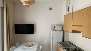 Cocina compacta con mesa de comedor, sillas blancas, televisor de pantalla plana, microondas, refrigerador y estufa a gas.